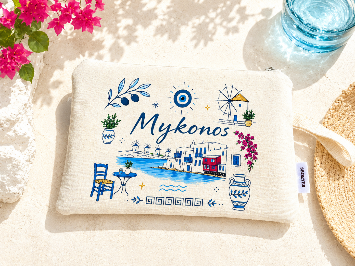 White pouch with 'Mykonos' design on a sandy background with flowers and a glass of water.