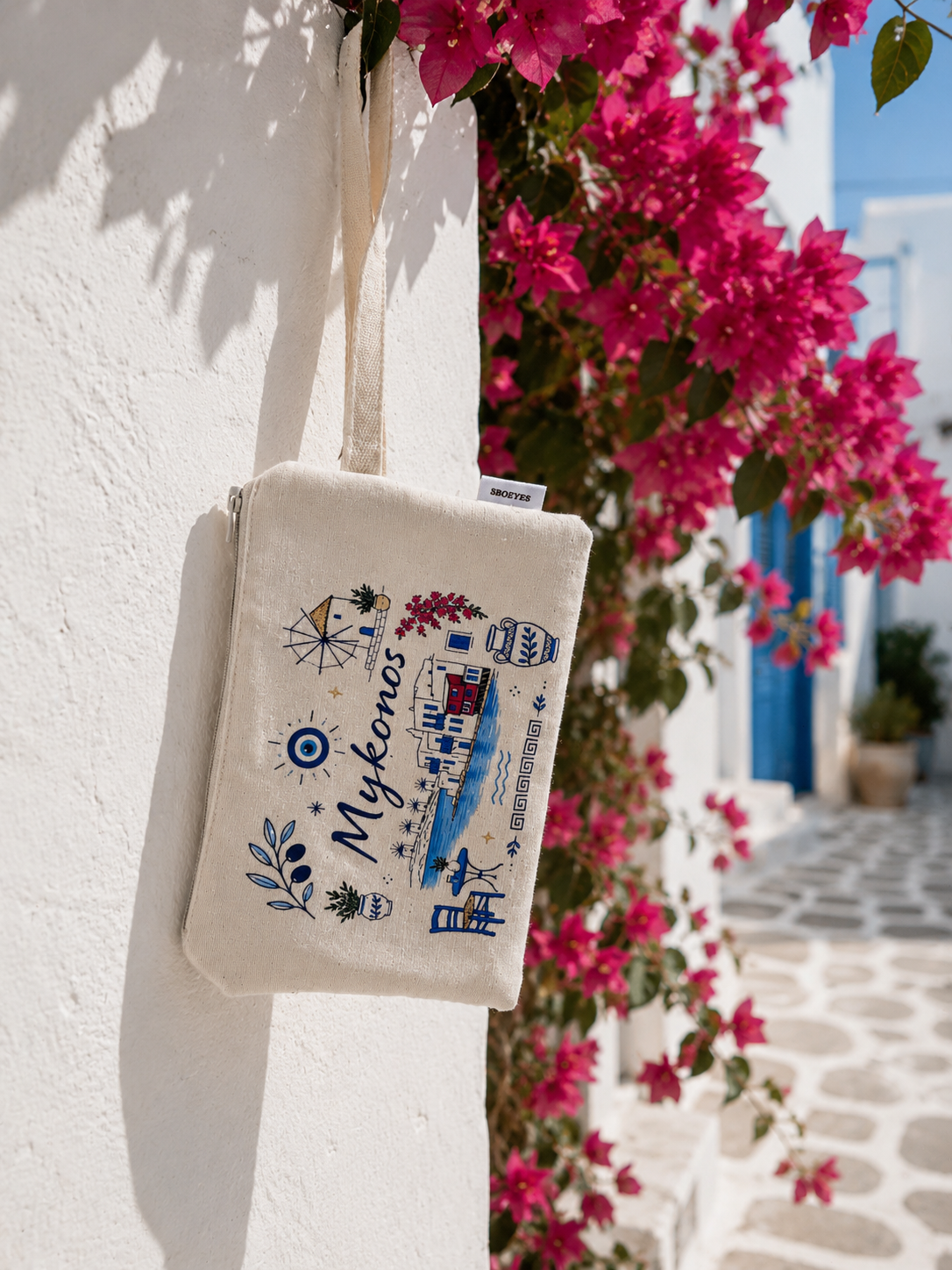 Tote bag with 'Mykonos' design hanging on a white wall with pink flowers in the background