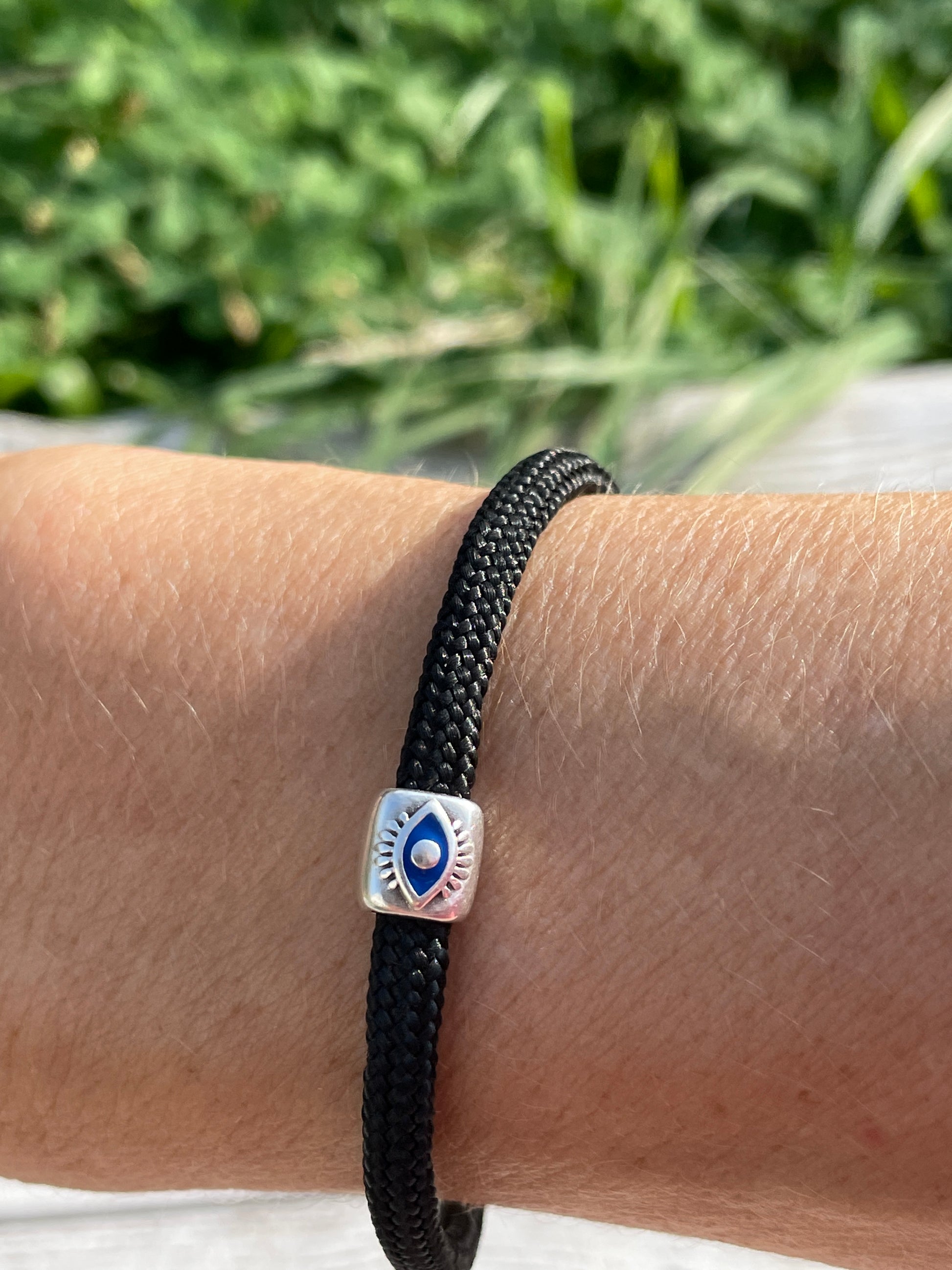 Black braided bracelet with a silver evil eye charm on a wrist against a blurred green background