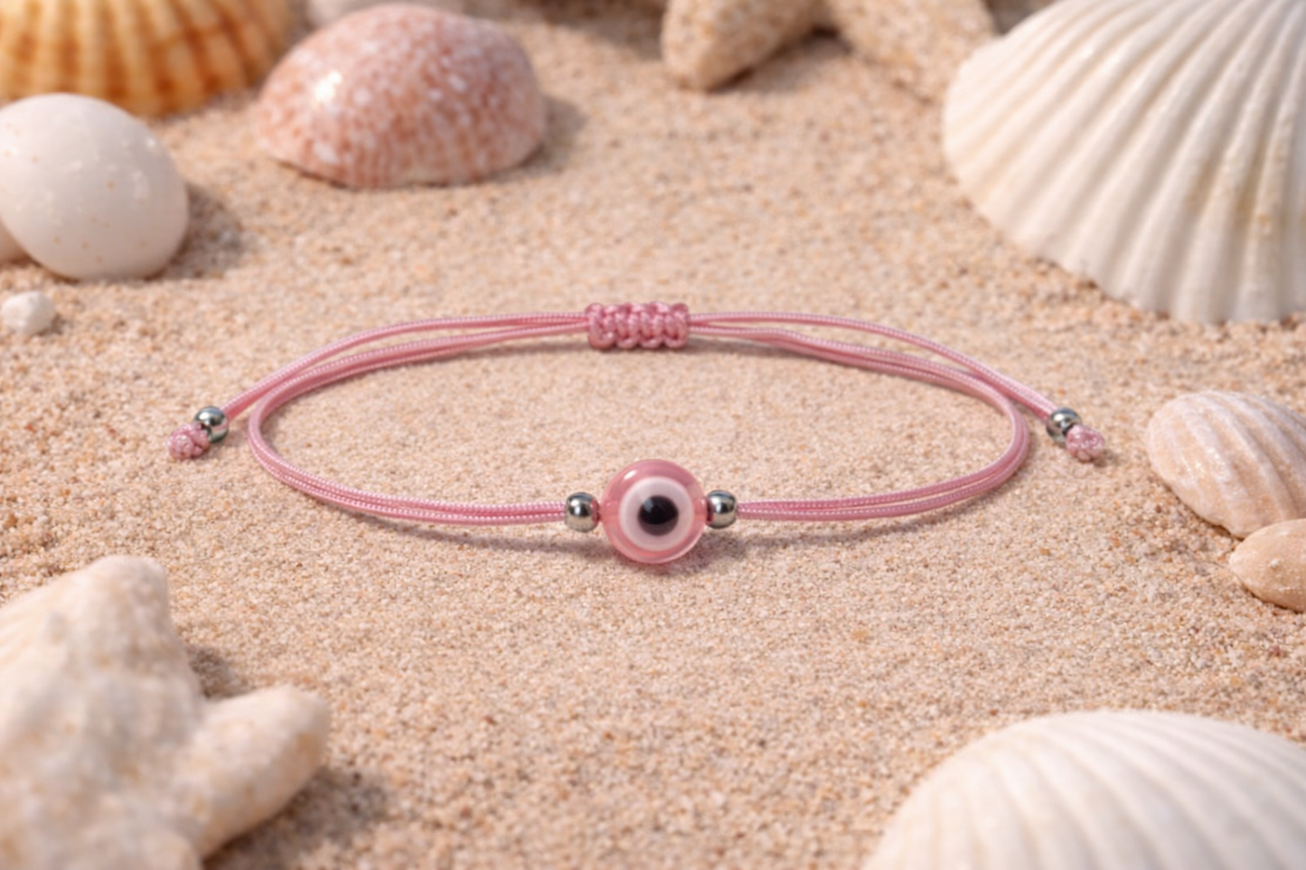 Pink bracelet with a black evil eye bead on sand surrounded by seashells