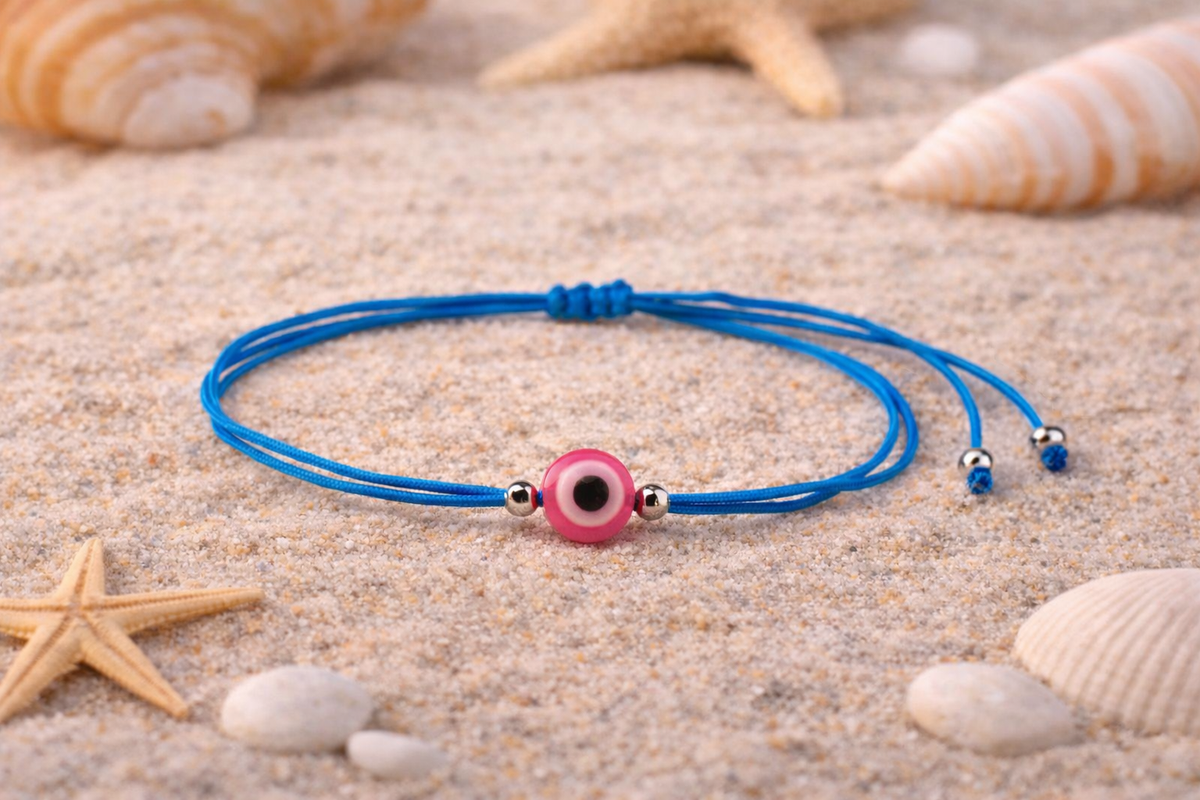 Blue bracelet with a pink evil eye charm on sand with starfish and shells