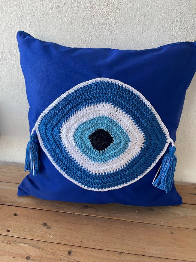 Blue pillow with a crochet evil eye design on a wooden surface.