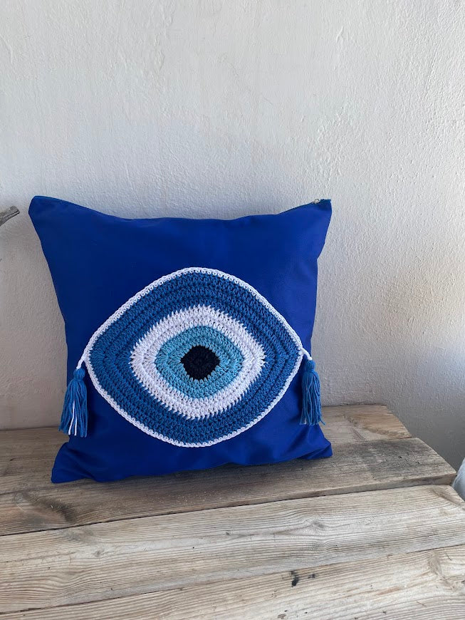 Blue pillow with a white embroidered eye design on a wooden surface.