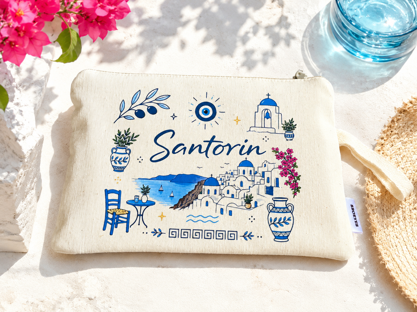 Beige pouch with Santorini design on a sandy background with flowers and water.