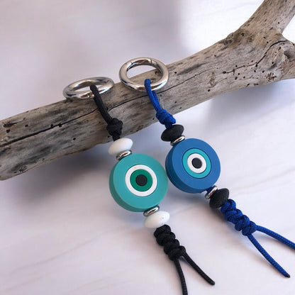Two silicone evil eye bag charms in blue and turquoise colors with black and white beads, attached to a nylon rope, displayed on a wooden branch.