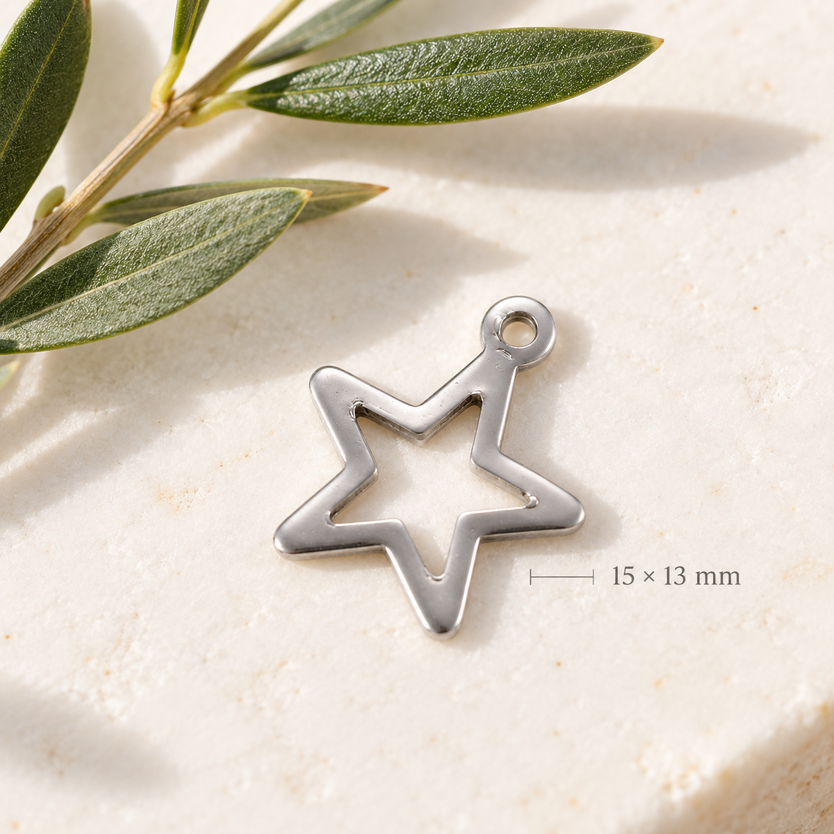 A silver open-star-shaped jewelry charm with a 15 x 13 mm size indicator overlay, resting on a textured beige stone surface next to green olive leaves.