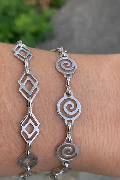 Silver bracelets with geometric and spiral designs on a wrist against a blurred natural background.