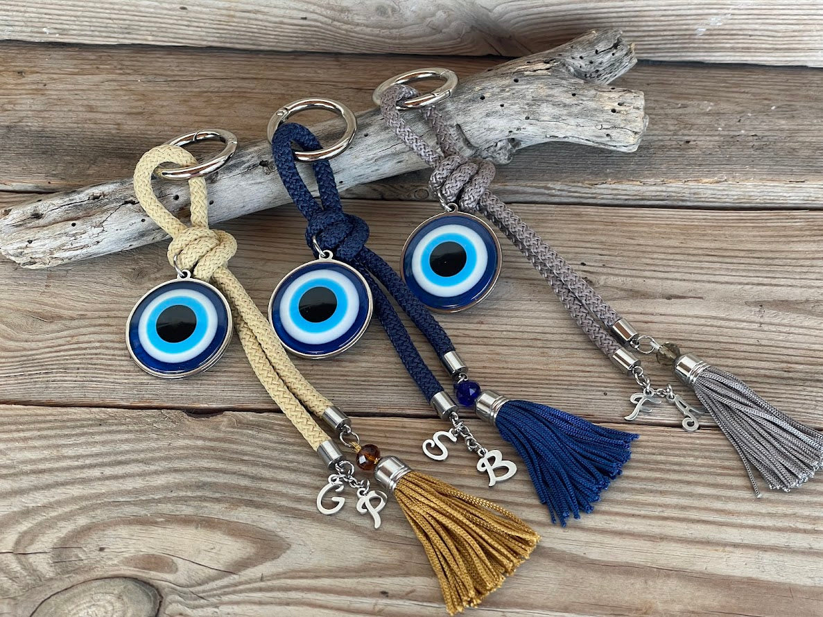 A collection of handcrafted evil eye bag charms with braided parachute cords and decorative tassels in dark blue, grey, and golden beige colors, displayed on a wooden surface.