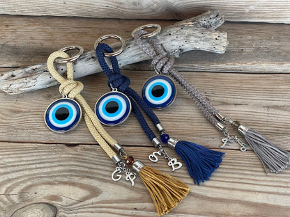 A collection of handcrafted evil eye bag charms with braided parachute cords and decorative tassels in dark blue, grey, and golden beige colors, displayed on a wooden surface.
