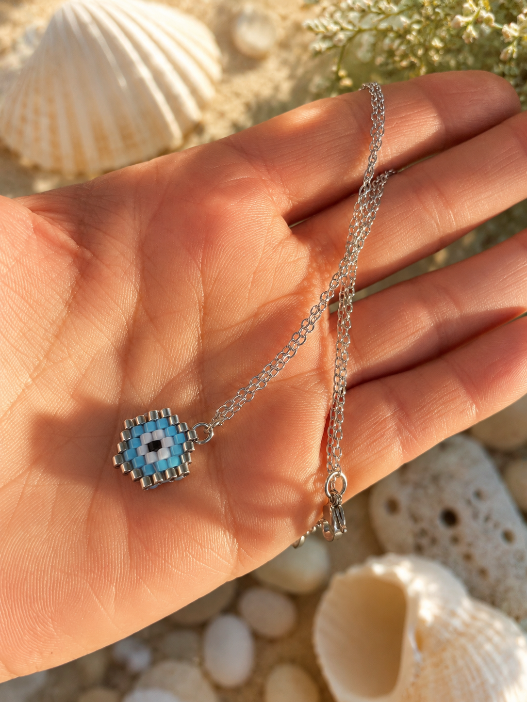 A silver chain necklace with a blue and white beaded evil eye pendant, held in a person's hand with seashells and sand in the background.