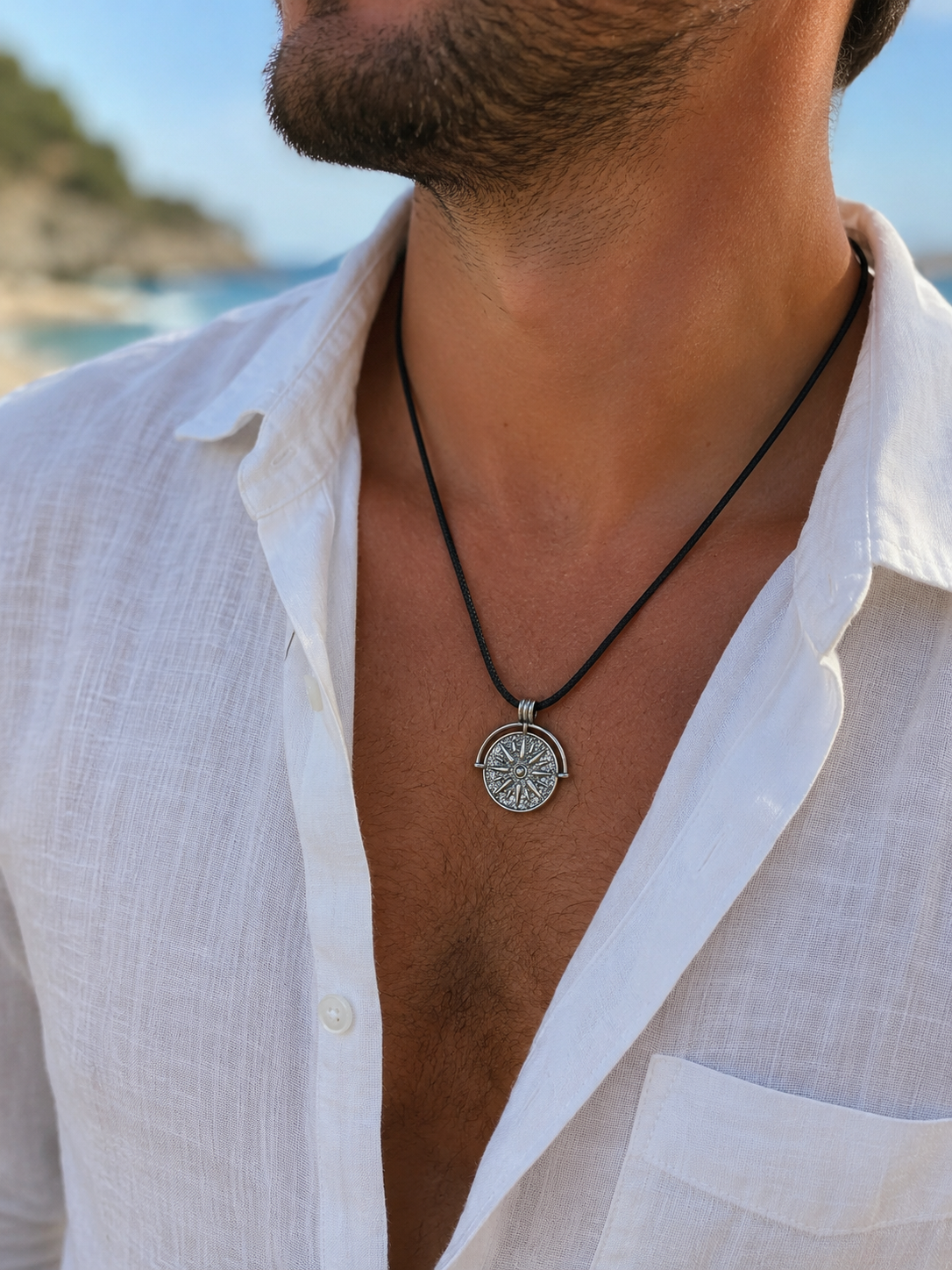 A close-up of a man wearing a silver compass pendant necklace on a black cord, styled with an open white linen shirt against a blurred beach background.