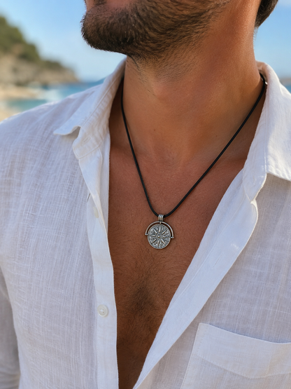 A close-up of a man wearing a silver compass pendant necklace on a black cord, styled with an open white linen shirt against a blurred beach background.