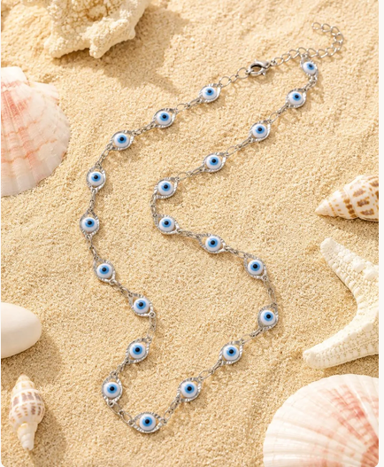 A silver chain necklace featuring multiple blue and white evil eye charms, styled on a sandy beach background with seashells and a starfish.