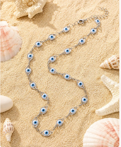 A silver chain necklace featuring multiple blue and white evil eye charms, styled on a sandy beach background with seashells and a starfish.