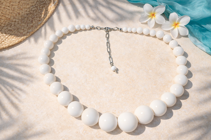 White beaded necklace on a sandy surface with flowers and a hat in the background