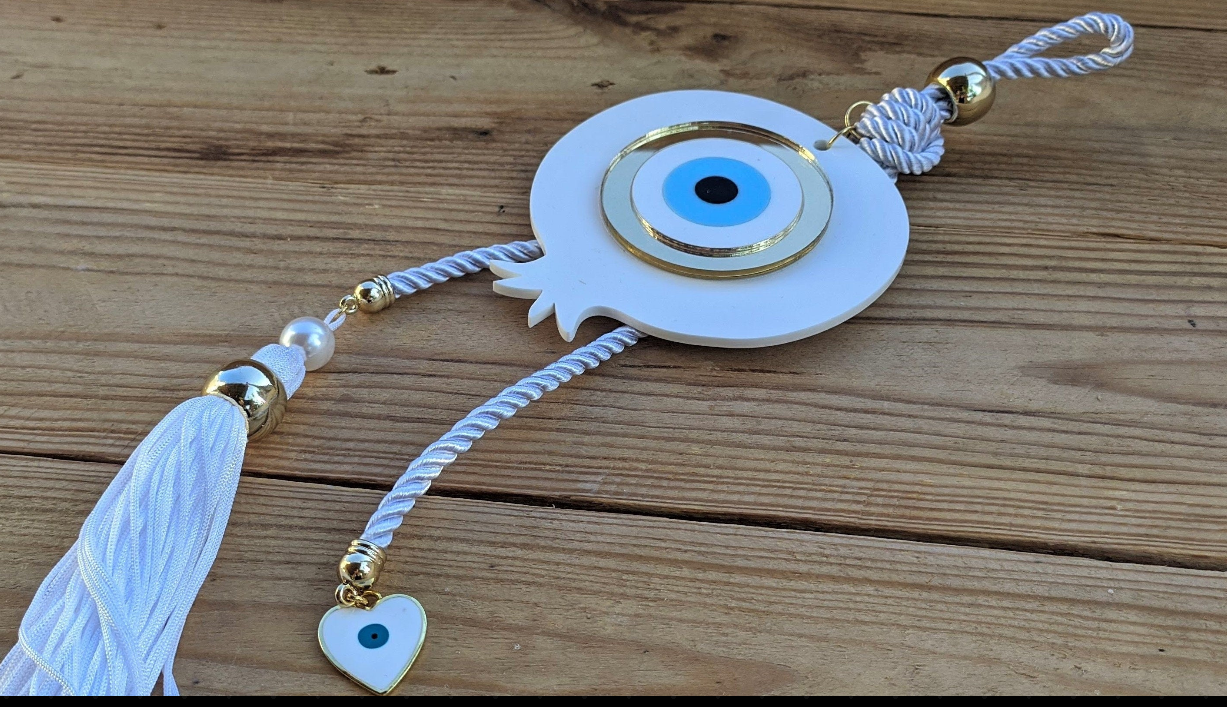 White plexiglass pomegranate wall hanging with a blue Greek evil eye, decorative tassel, and heart charm, symbolizing good luck and prosperity.