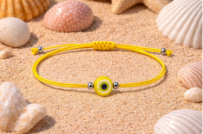 Yellow bracelet with a eye charm on sand surrounded by seashells