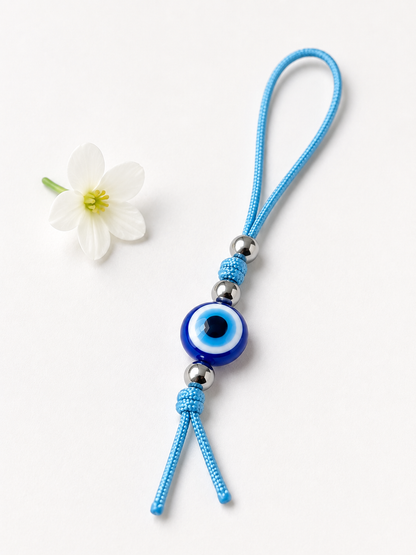 A blue knotted cord charm with a blue and white evil eye bead and silver metal beads, displayed on a clean white background next to a small white flower.