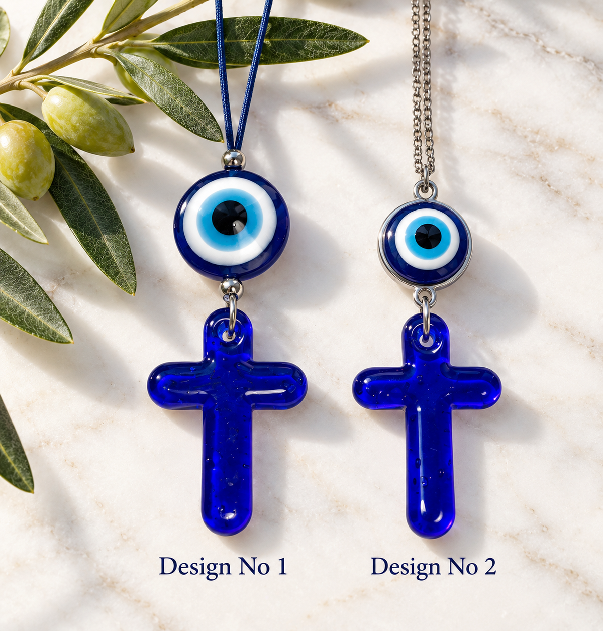 Two cobalt blue glass evil eye necklace designs displayed on a light marble surface with olive branches. Design No 1 has a blue cord and a blue glass cross pendant, while Design No 2 has a silver chain and a matching blue glass cross pendant.
