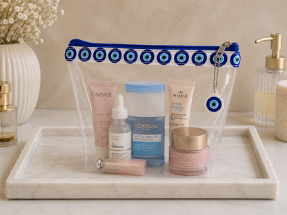 Clear PVC cosmetic bag with Greek evil eye trim and charm, filled with beauty products.