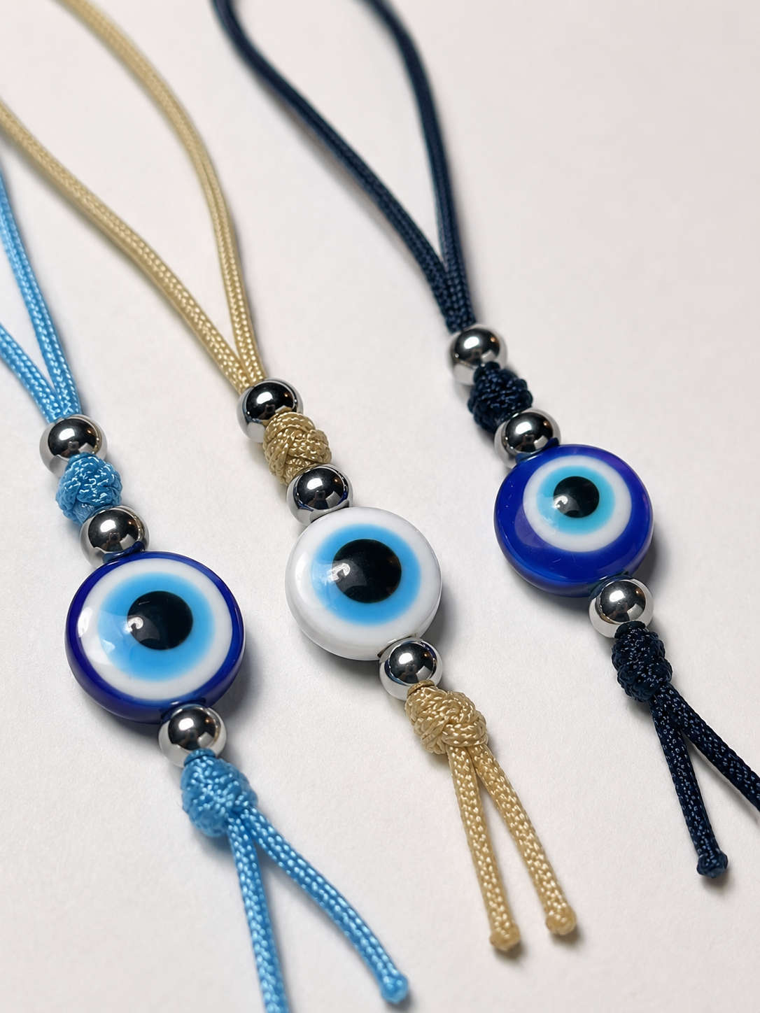 Three cord necklaces with evil eye beads and silver beads, arranged diagonally on a light gray background. One necklace is light blue, one is beige, and one is dark navy blue.