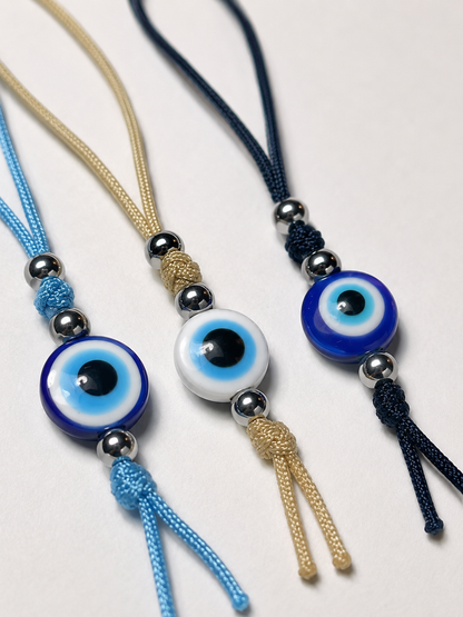 Three cord necklaces with evil eye beads and silver beads, arranged diagonally on a light gray background. One necklace is light blue, one is beige, and one is dark navy blue.