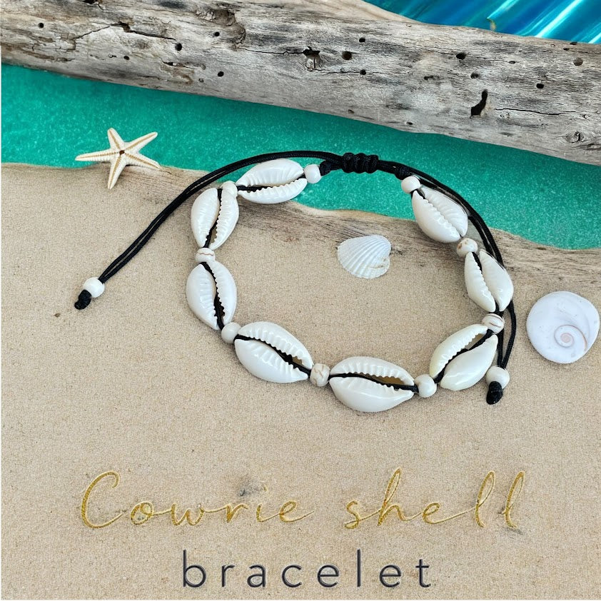 Cowrie shell bracelet on a sandy surface with driftwood and shells in the background.