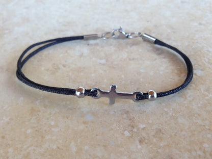 Black cord bracelet with silver clasp and cross charm on a beige surface