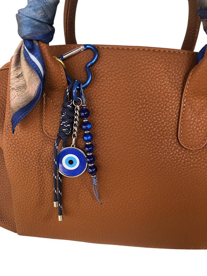 A blue and brown handbag with a stainless steel evil eye charm and beaded decoration, attached to a leather strap.
