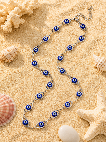 A silver chain necklace featuring multiple blue and white evil eye charms, arranged in a wave pattern on a sandy beach background with seashells and coral.