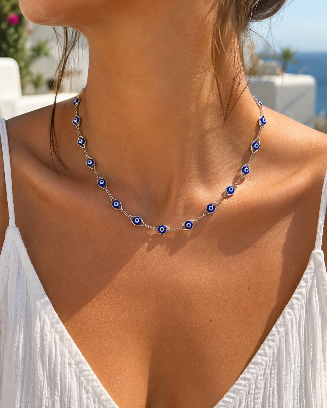 A close-up of a woman wearing a silver chain necklace adorned with blue and white evil eye charms, set against a bright outdoor background.