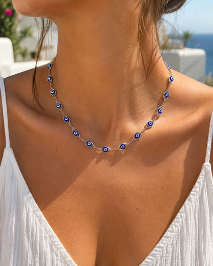 A close-up of a woman wearing a silver chain necklace adorned with blue and white evil eye charms, set against a bright outdoor background.