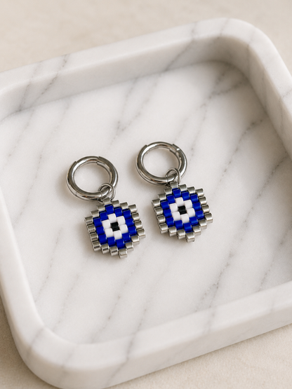A pair of silver-toned hoop earrings with square evil eye charms in blue, white, and black, displayed on a white marble tray.