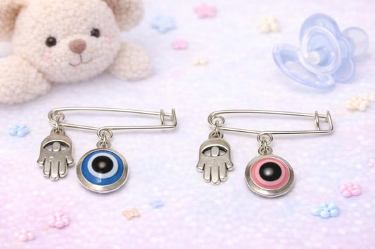 Two decorative pins with hand and eye designs on a soft pink background with toys and pacifiers.