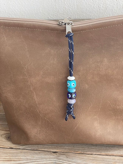A beaded bag charm with multicolored beads and a resin evil eye pendant, attached to a nylon cord on a brown leather bag.