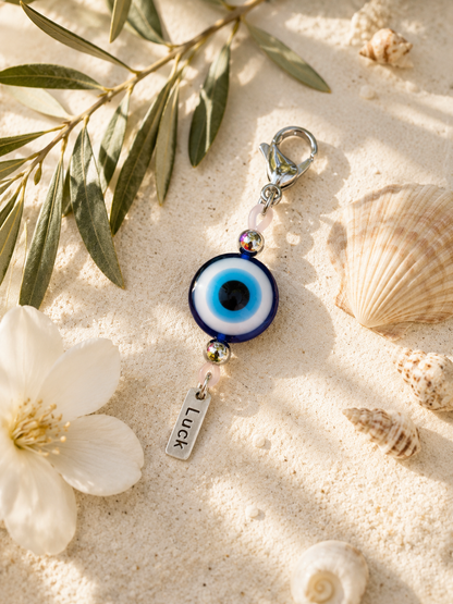 A beaded keychain or bag charm laid on sandy ground with seashells, olive branches, and a white flower. The charm has a blue-and-white evil eye bead, silver-toned metal beads, a lobster clasp, and a small rectangular metal tag engraved with the word "Lucky.