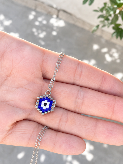 A silver chain necklace with a blue and white beaded evil eye pendant, held in the palm of a hand against an outdoor background.