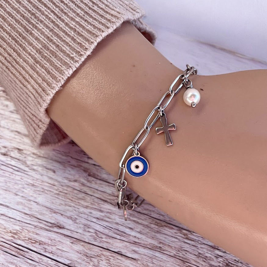 A stainless steel bracelet with multiple charms including an evil eye, a heart, and a cross, worn on a wrist.