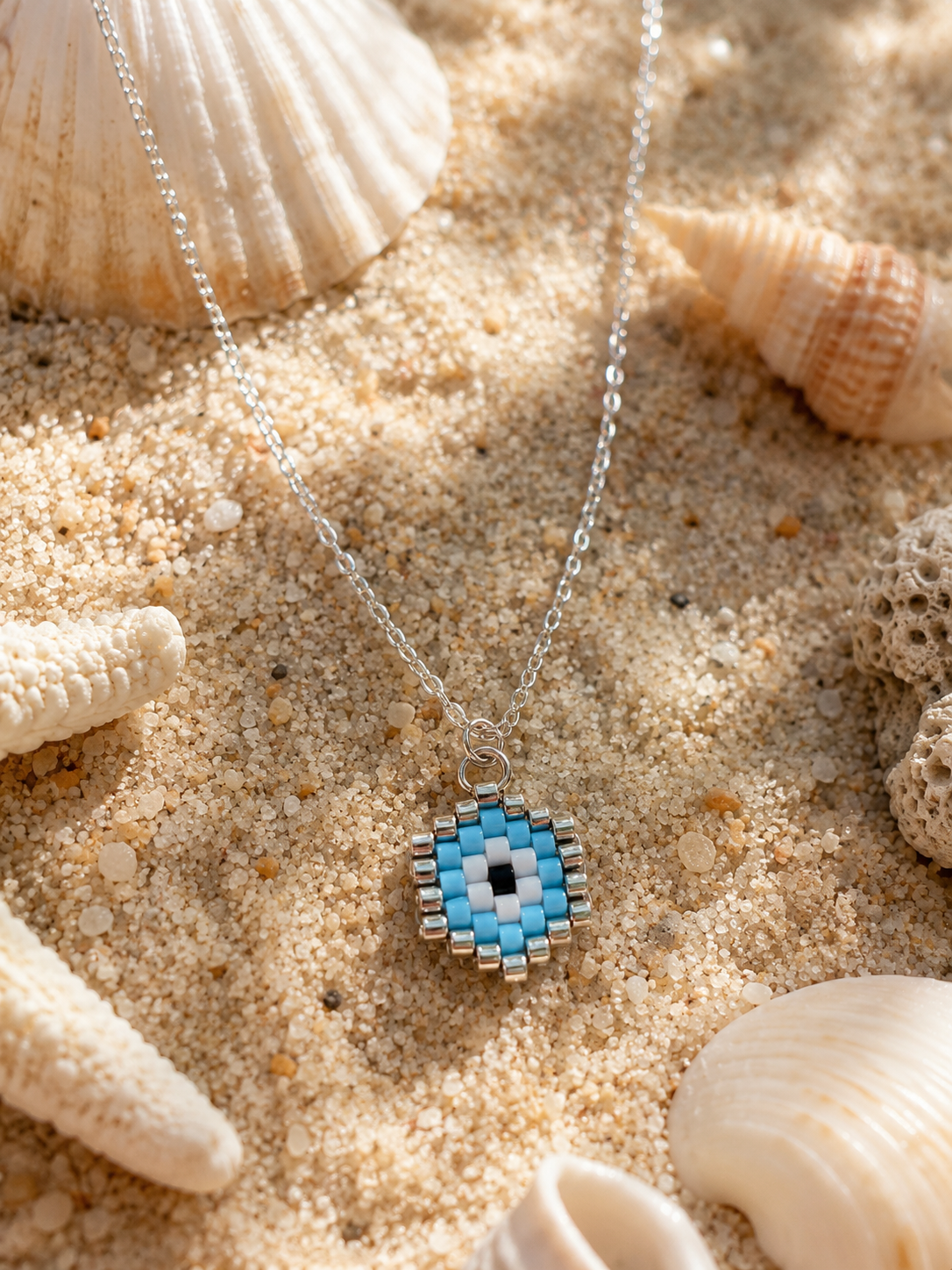 A silver chain necklace with a beaded circular pendant featuring a blue, white, and black evil eye design, styled on sand with seashells.