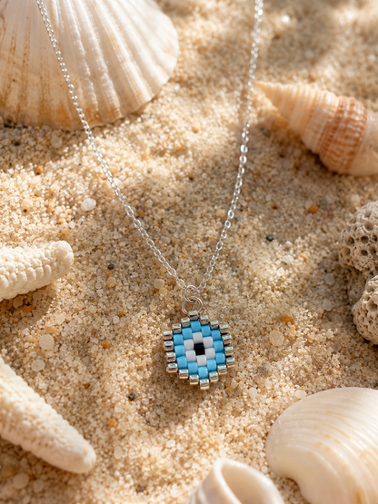 A silver chain necklace with a beaded circular pendant featuring a blue, white, and black evil eye design, styled on sand with seashells.