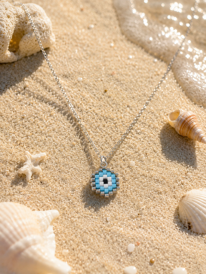 A silver chain necklace with a beaded circular evil eye pendant in shades of blue, white, and black, styled on sandy beach with seashells and ocean foam.