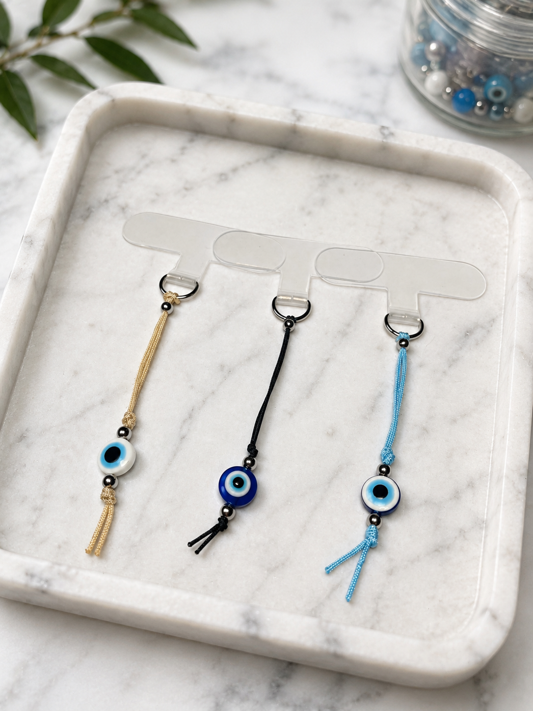 Three corded evil eye keychains in gold, black, and blue, displayed on a clear plastic hanger over a white marble tray.