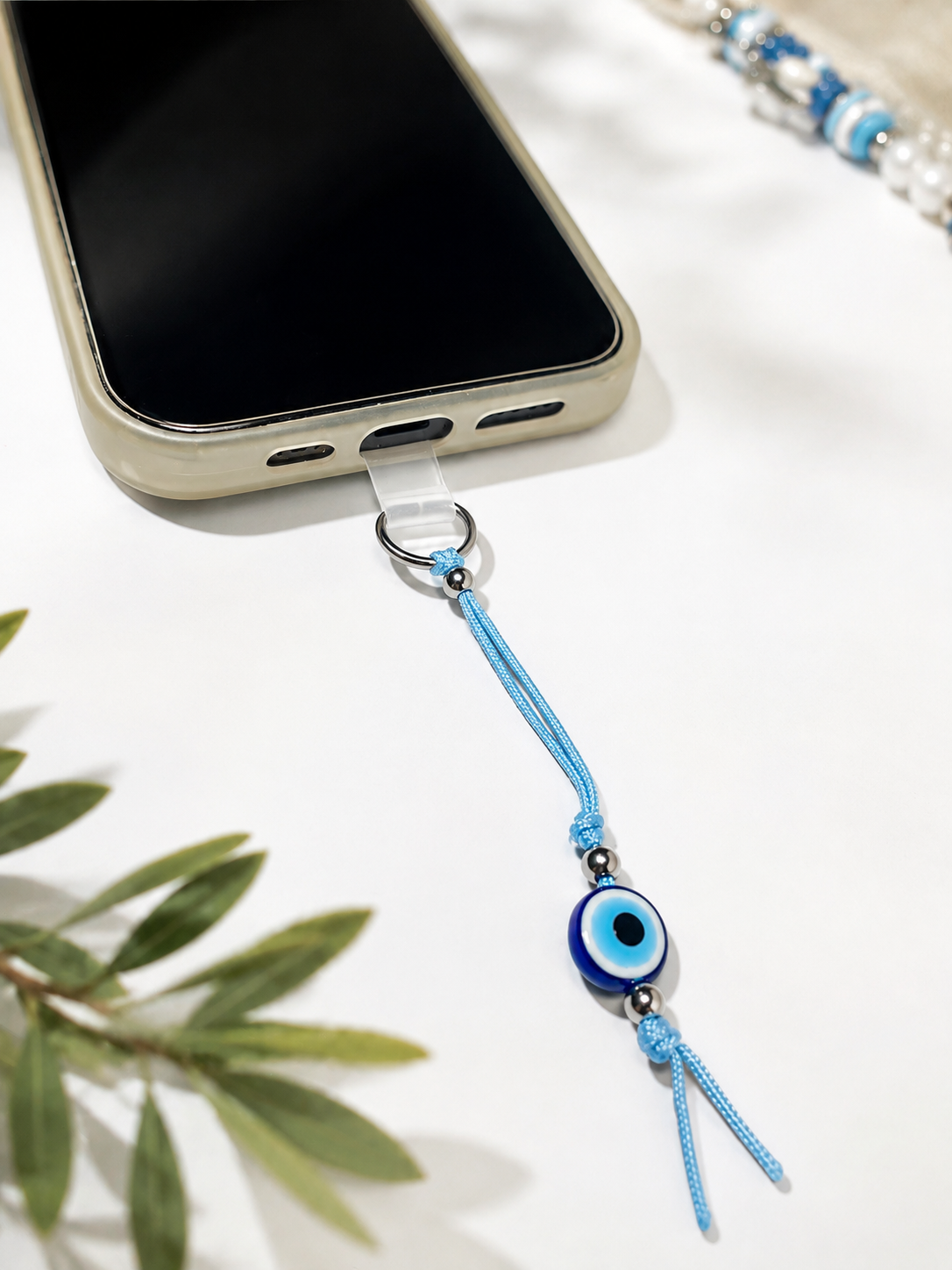 A light blue cord phone strap with a blue and white evil eye bead and silver metal hardware, shown attached to a smartphone on a white surface with decorative green leaves.
