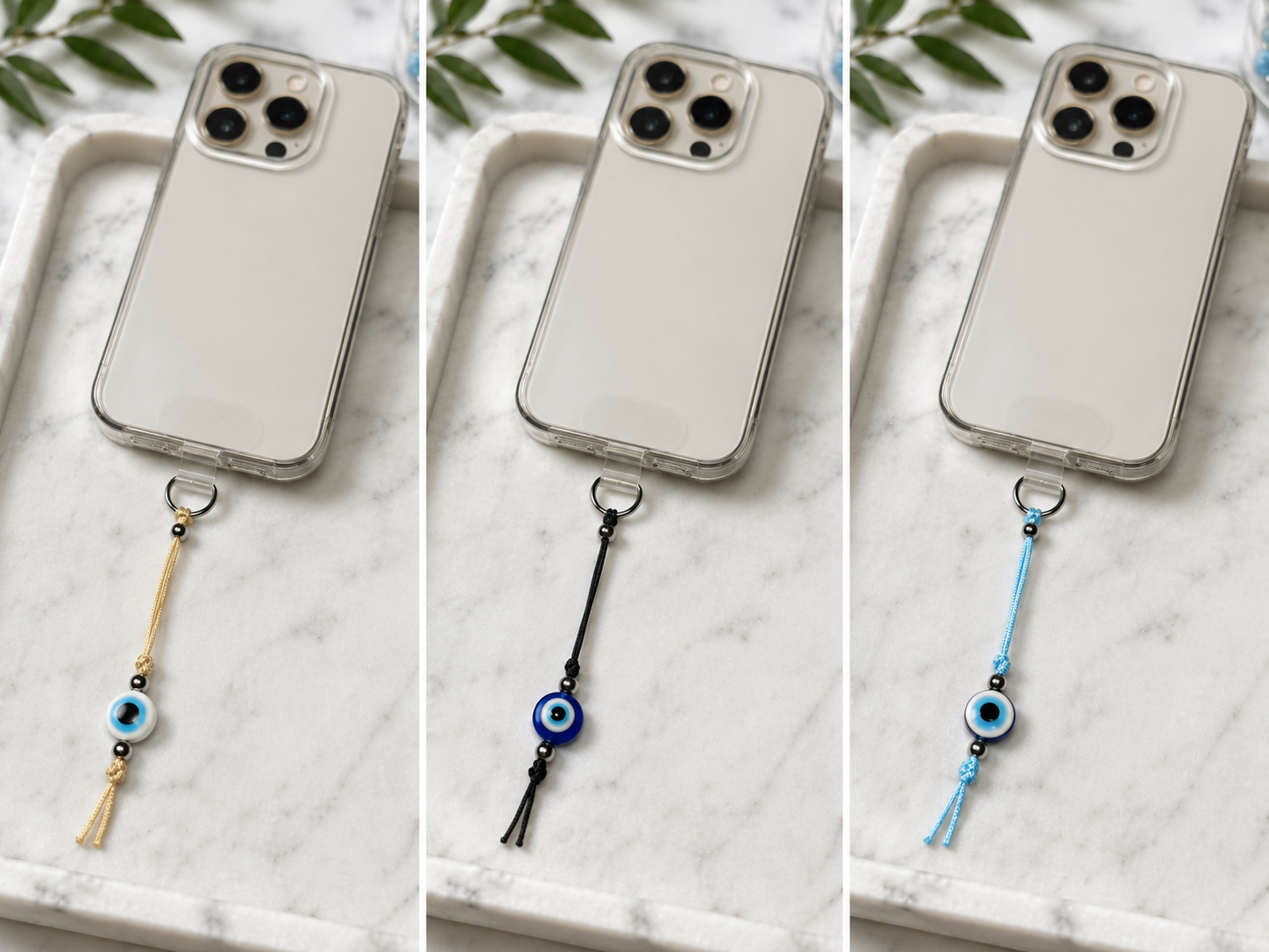 Three phone cases shown side by side on a white marble tray, each with a dangling cord charm featuring an evil eye bead and tassel. The left case has a gold cord, the middle has a black cord, and the right has a light blue cord.