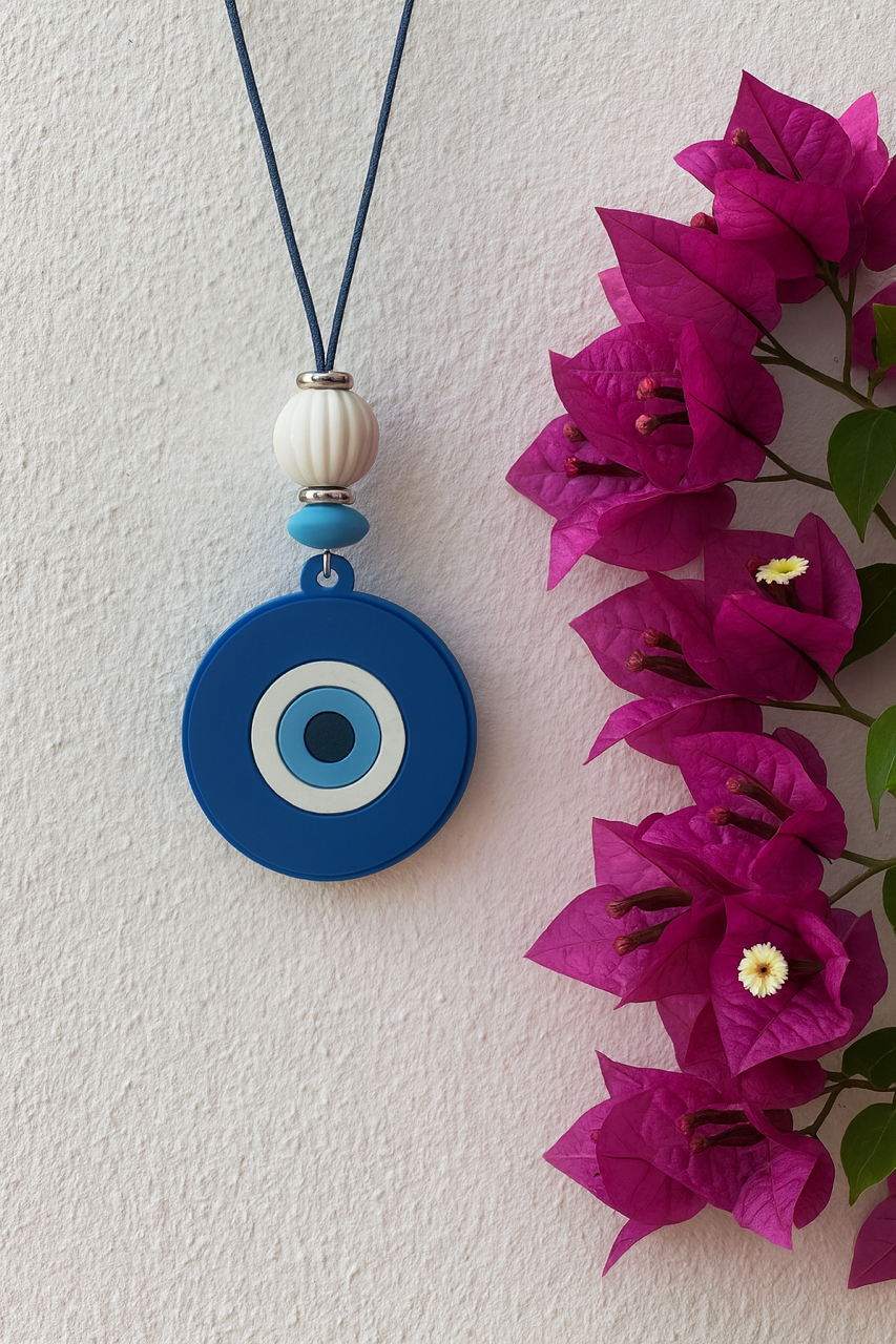 Blue evil eye pendant necklace with a white bead on a string, next to pink flowers against a light background.