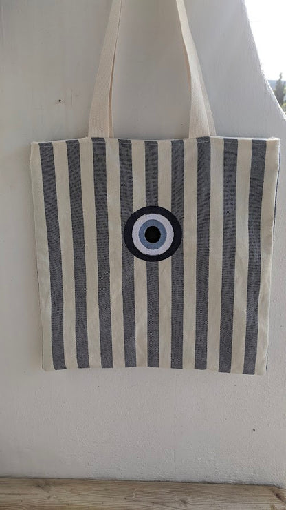 A striped canvas tote bag with turquoise stripes and a central embroidered evil eye design in blue.