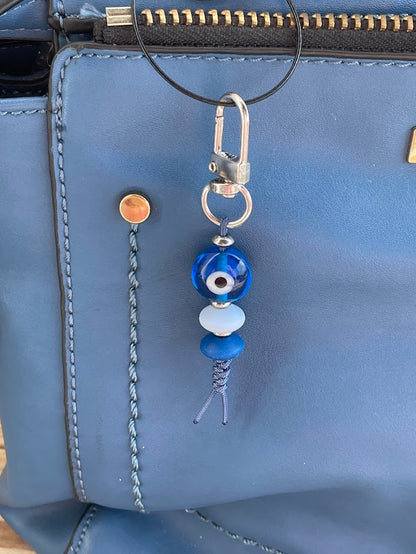 A blue glass evil eye bag charm with a silver tone snap clasp, displayed in front of a brown handbag.