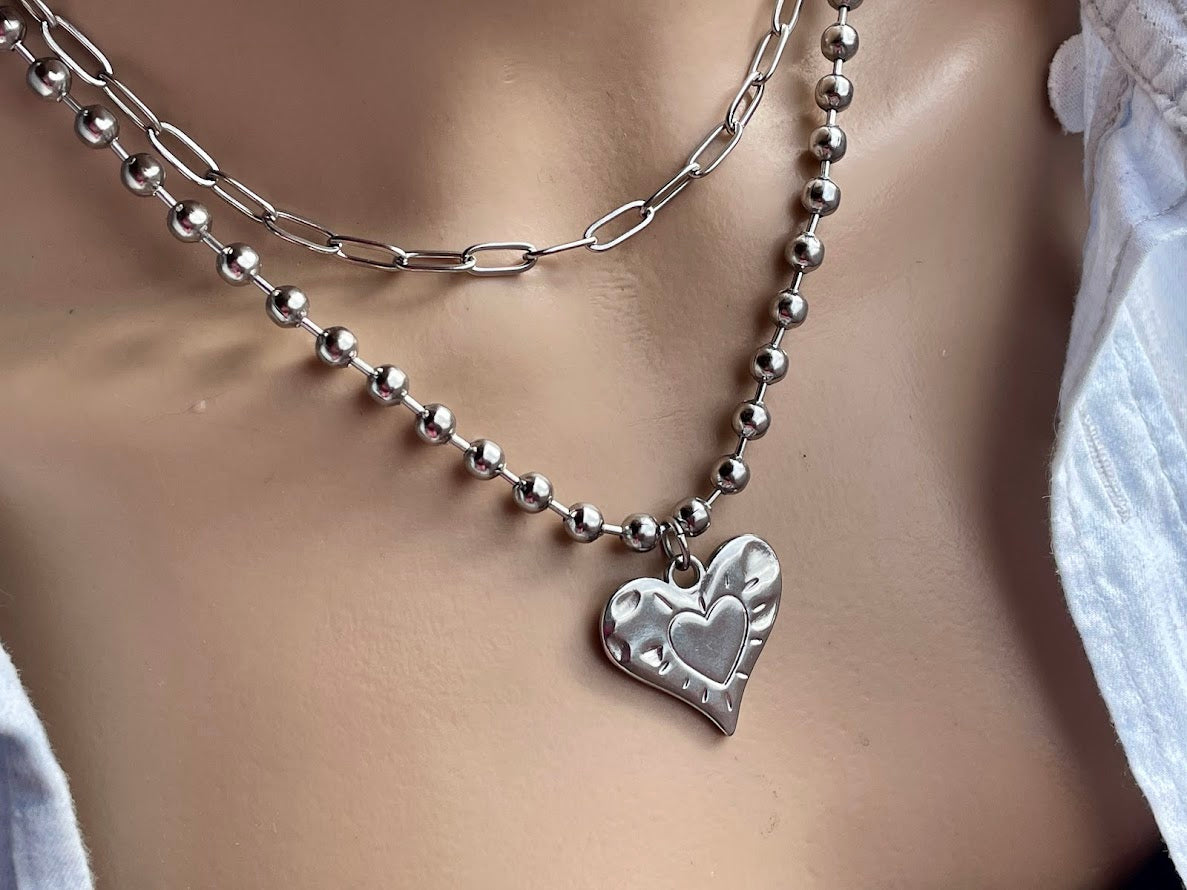 A silver stainless steel heart charm necklace with a ball chain on a mannequin.