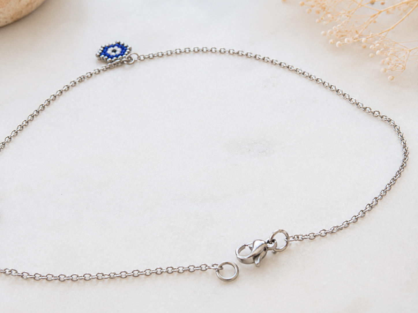 A delicate silver chain bracelet with a small blue and white evil eye charm, displayed on a light textured surface.