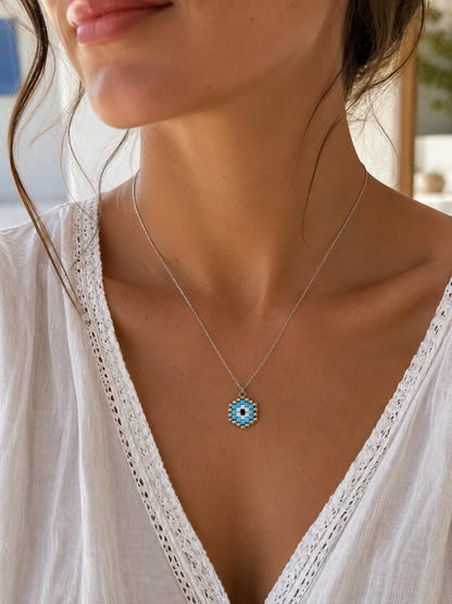 A close-up of a woman wearing a delicate silver chain necklace with a blue and white evil eye pendant, paired with a white lace-trimmed top.